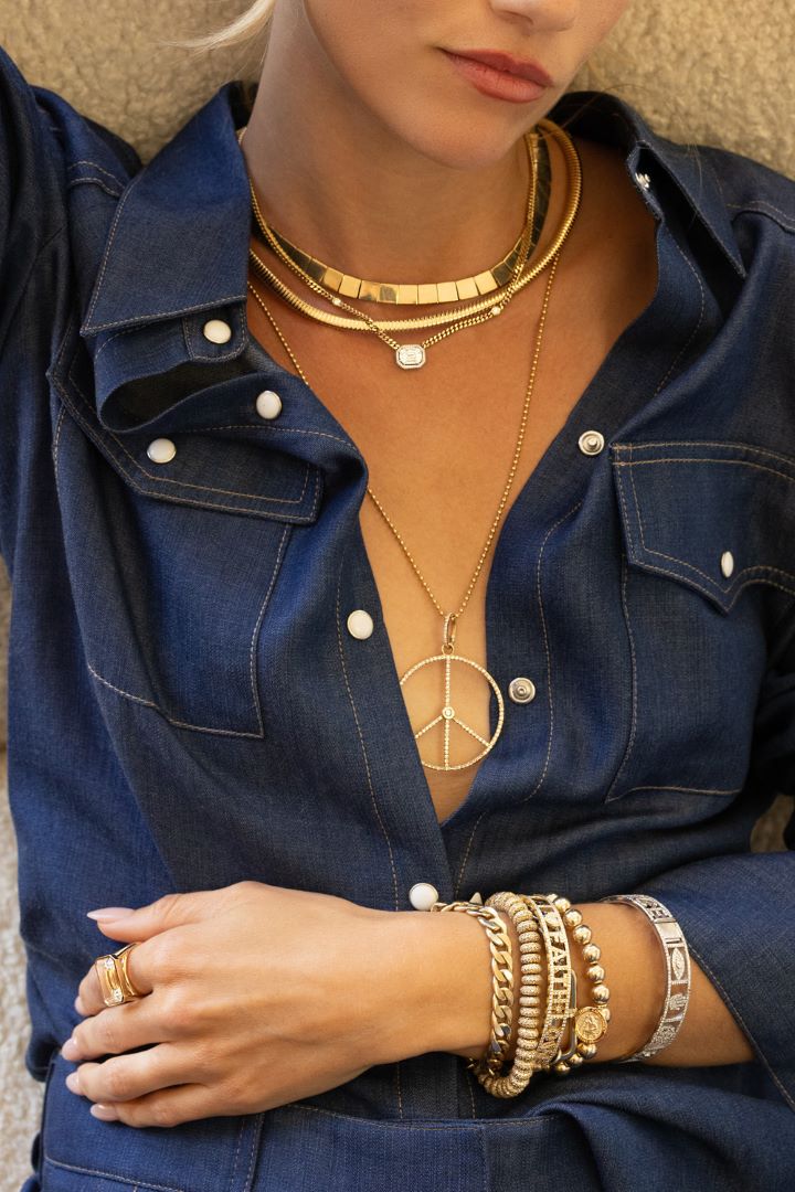 Layers of 14k gold Sheryl Lowe necklaces and a stack of 14k gold Sheryl Lowe bracelets pictured on a woman in a denim top.